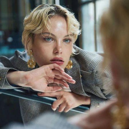 Model with blonde hair and ornate earrings wearing a grey plaid blazer, reflected in a mirror.