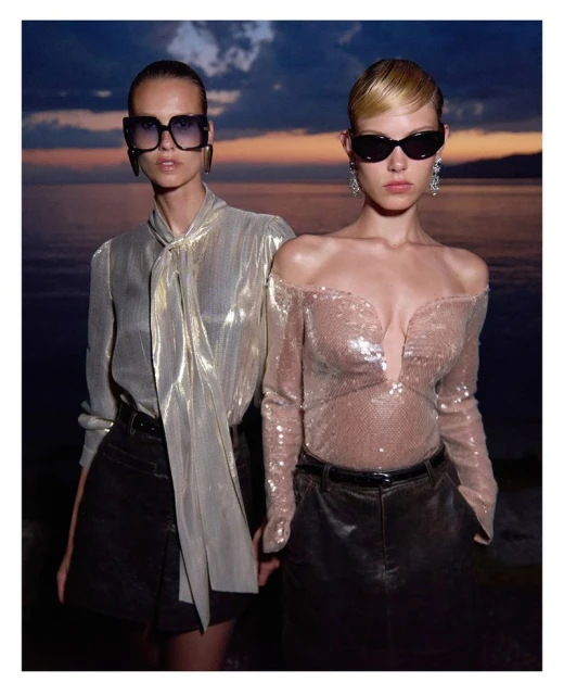 Two women pose outdoors at dusk wearing sunglasses, shiny blouses, and dark mini skirts. The ocean and colorful sky appear in the background.