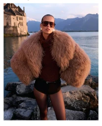 Model wearing a fluffy textured coat and sunglasses posing near a lake and stone building for Self-Portrait.