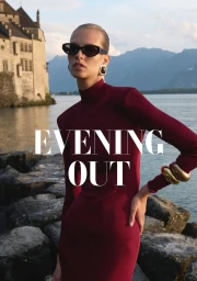 Model wearing a burgundy high-neck dress and dark sunglasses posing outdoors by a lake with a stone castle and mountains