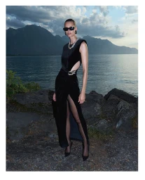 Model wearing a black velvet dress and sunglasses standing by a lake with mountains in the background