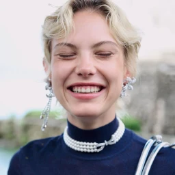 Close-up portrait of a smiling woman with short blonde hair wearing a dark blue turtleneck and crystal earrings.