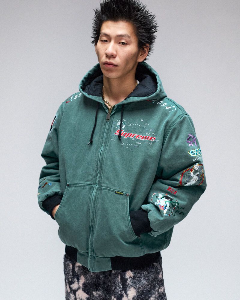 Asian man in a teal Supreme hooded jacket with graphics. He has spiky hair and is wearing a gold chain against a neutral background.