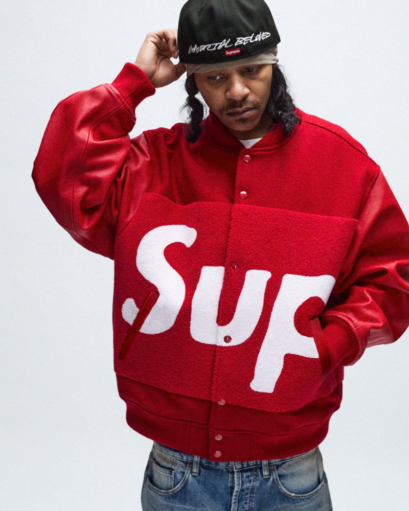 Adult male wearing a red Supreme wool jacket, black cap and blue jeans in a studio setting, posing for a fashion portrait.