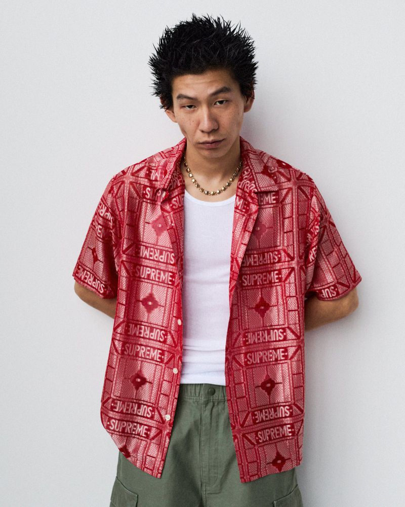 Asian man in a red patterned Supreme shirt, white undershirt, and green pants poses against a white wall in a studio-style fashion portrait.