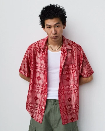 Asian man in a red patterned Supreme shirt, white undershirt, and green pants poses against a white wall in a studio-style fashion portrait.