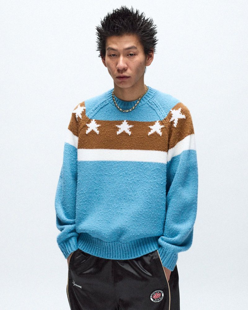 Portrait of a young adult man wearing a star-patterned sweater and black track pants posing in front of a white background.