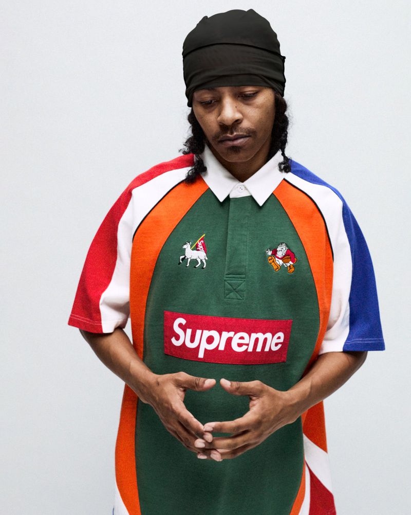Portrait of an adult male wearing a green collared shirt with a Supreme logo and a black head covering, against a white background.