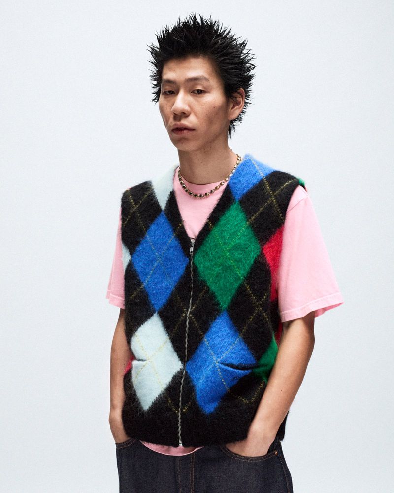 Adult male poses in a studio, wearing a colorful argyle vest over a pink tee, and dark wash jeans against a white background.