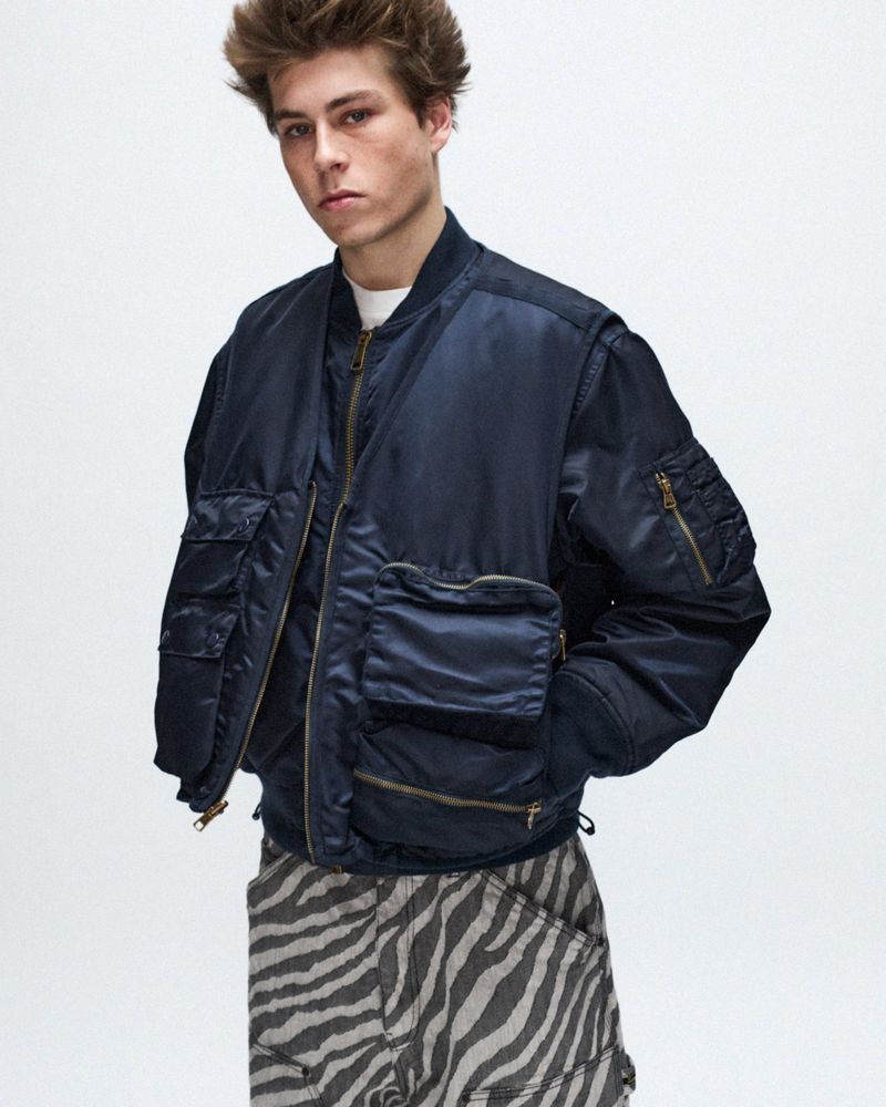 Male fashion model posing in an unbuttoned navy bomber jacket with many pockets and zebra-print pants in a neutral studio.