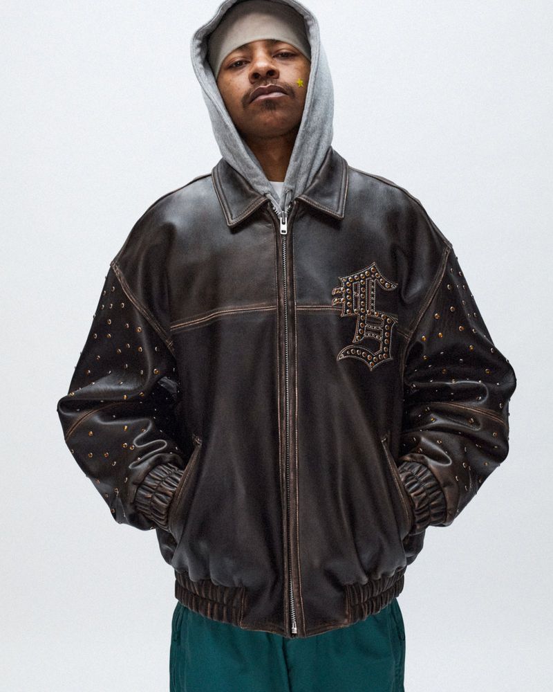 An adult male poses indoors wearing a gray hooded sweatshirt and dark brown studded leather jacket, showcasing fashion and apparel.