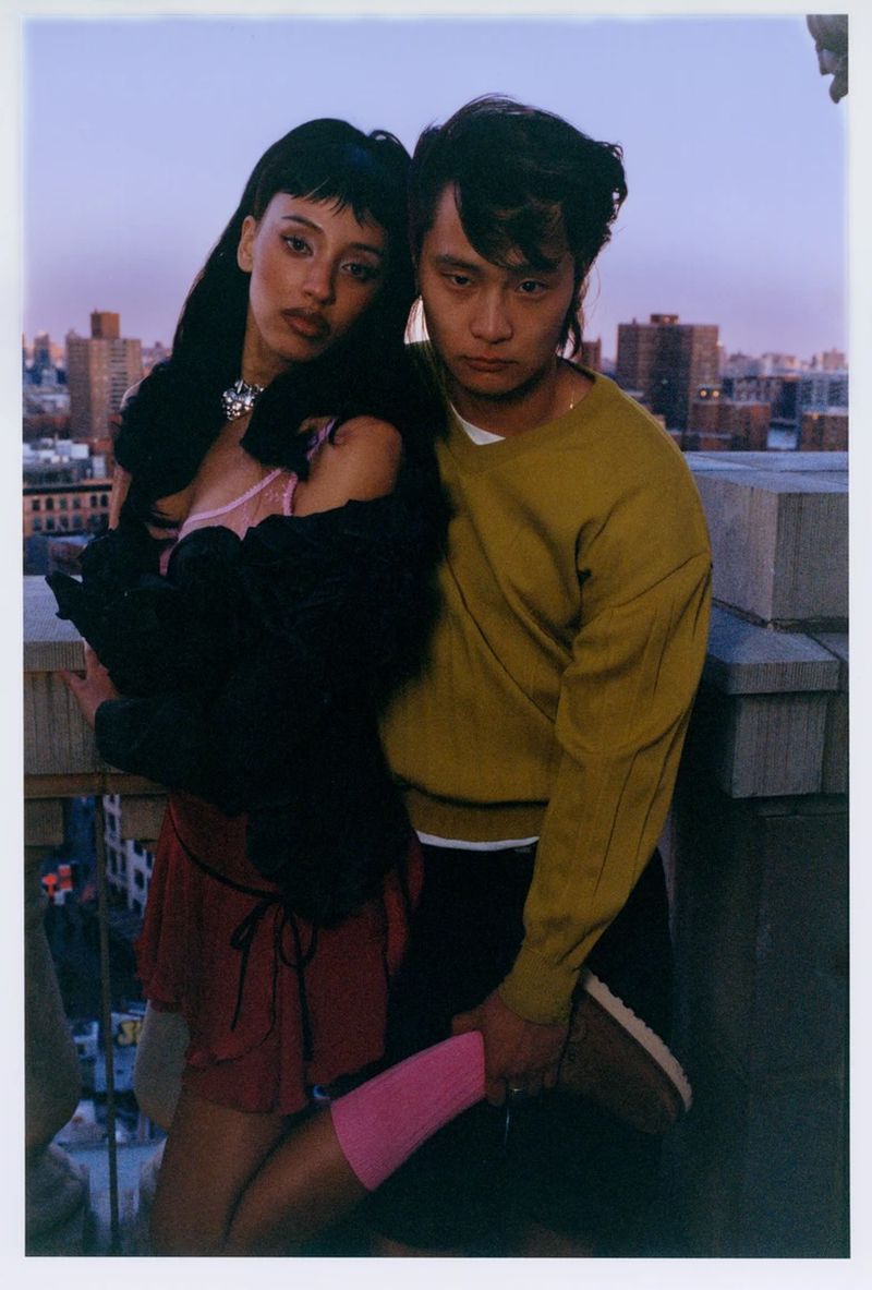 Candid shot of a stylish couple posing on a rooftop in an urban setting with city buildings and a vibrant dusky sky.