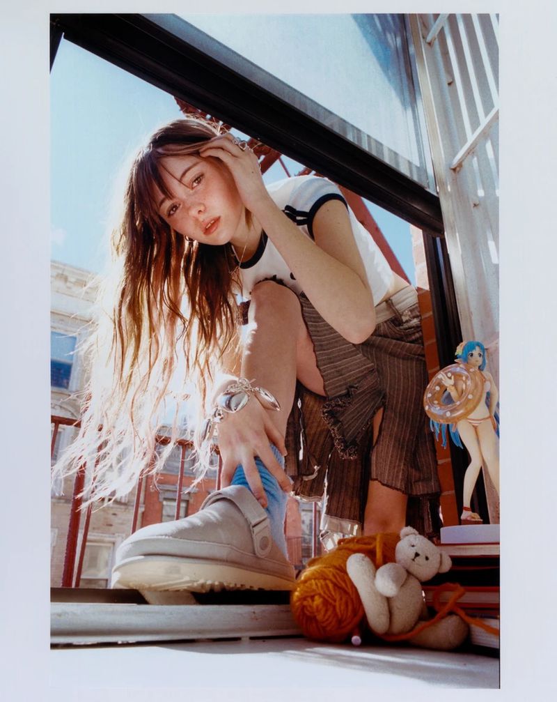 A young woman in trendy clothing poses with a teddy bear, yarn, and an anime figurine on a balcony with the sky in the background.