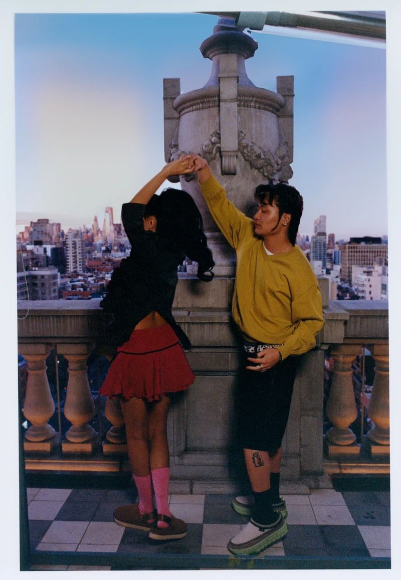 A young couple poses on a balcony with a city skyline at dusk. The woman wears a black top and pink skirt, while the man wears a yellow sweater.