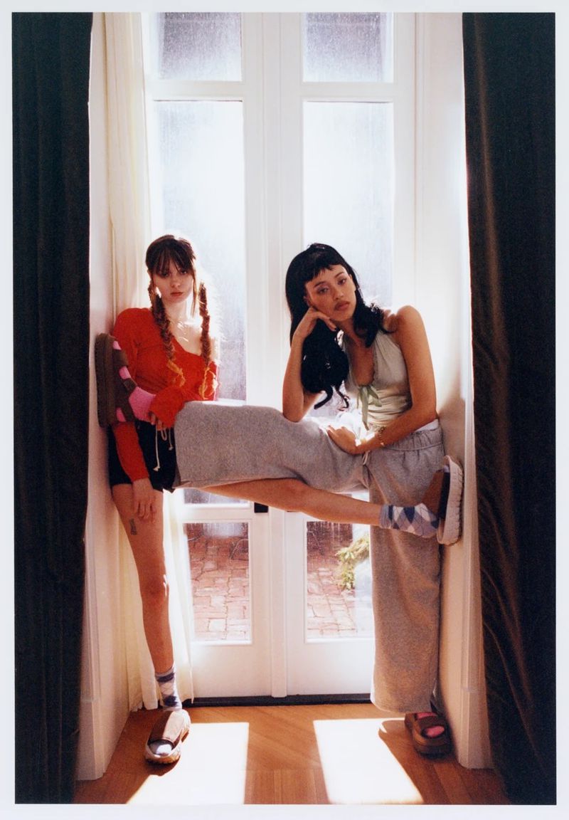 Two women in a window. One is seated on the sill wearing a grey skirt, socks, and platform sandals. The other stands next to her.