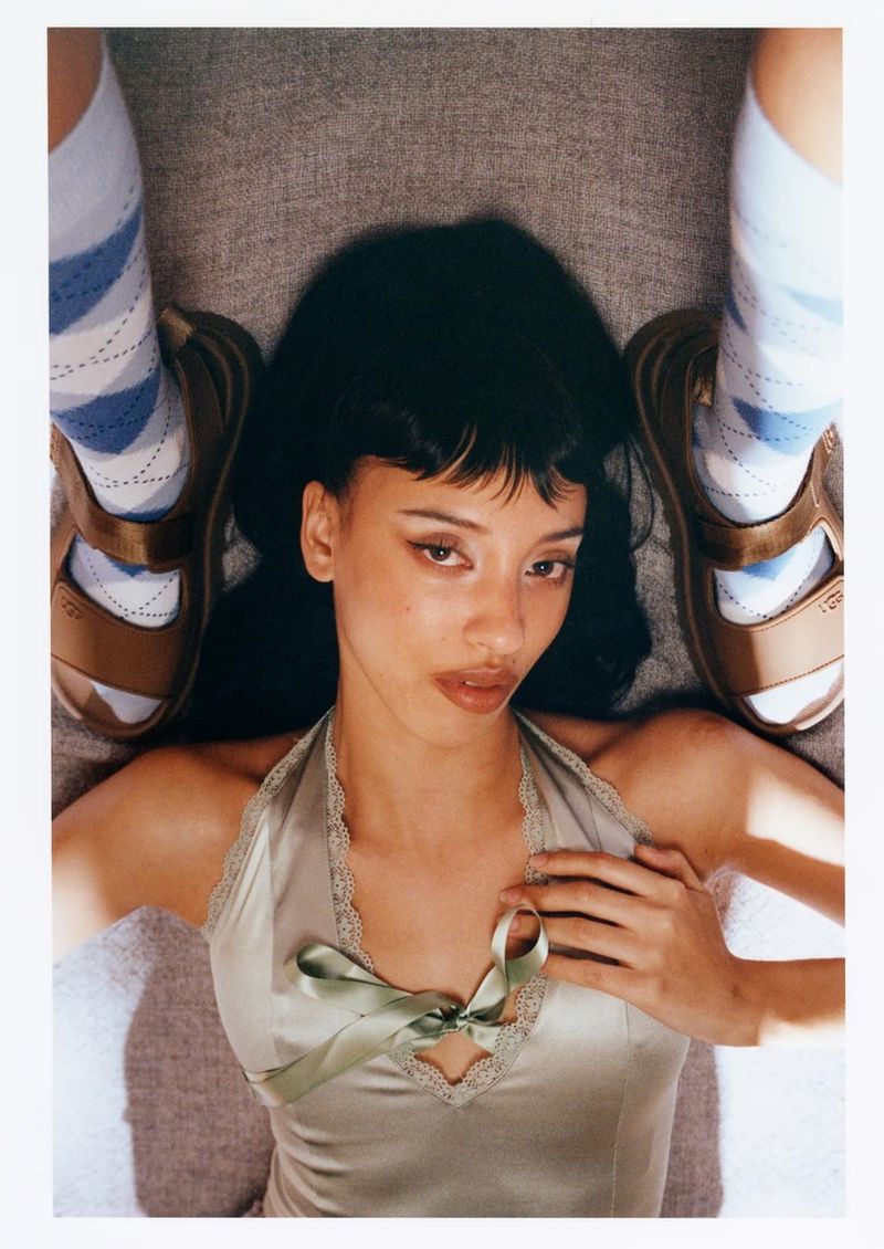 A young woman with dark hair poses with sandals and argyle socks, shot from a high angle on a neutral grey background, creating a Y2K aesthetic.