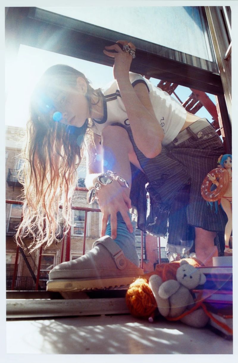 Young adult woman poses on a balcony with bright sunlight, wearing a white top, striped skirt, and a large shoe with visible outdoor urban background.