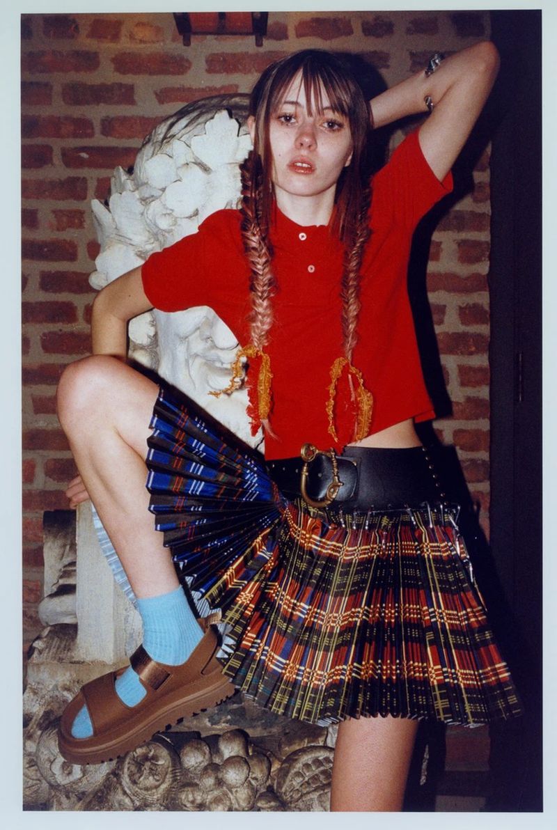 Young woman in red top and plaid skirt poses with ornate sculpture. She wears pigtails with colorful beaded accents. Indoor brick wall background.