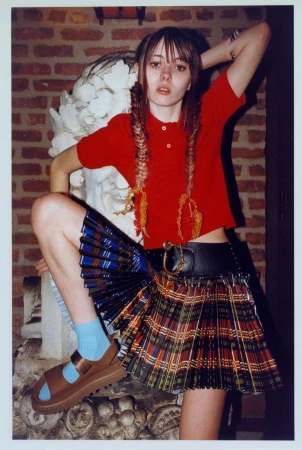 Young woman in red top and plaid skirt poses with ornate sculpture. She wears pigtails with colorful beaded accents. Indoor brick wall background.