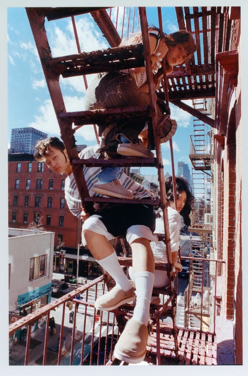 Three young adults pose on a rusty exterior fire escape of an urban building under a partly cloudy sky, adding a touch of youthful energy.
