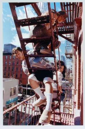 Three young adults pose on a rusty exterior fire escape of an urban building under a partly cloudy sky, adding a touch of youthful energy.