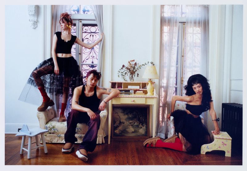 Three fashion models pose indoors with furniture in a brightly lit room; a female model stands by a window while the other two sit.