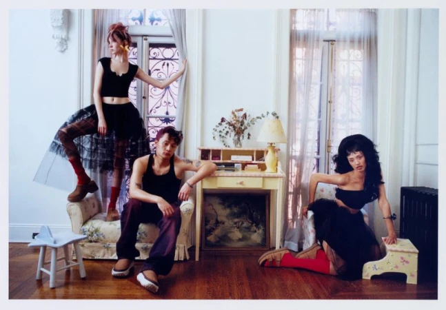 Three fashion models pose indoors with furniture in a brightly lit room; a female model stands by a window while the other two sit.