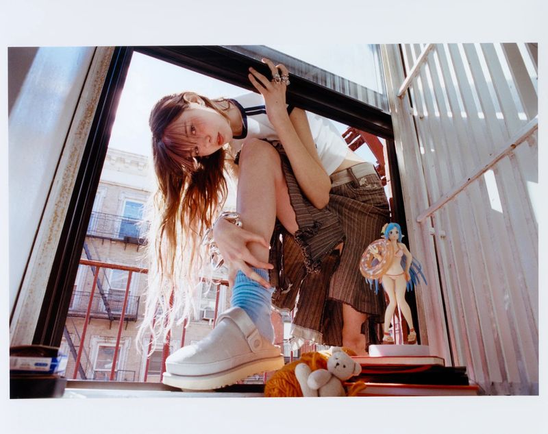 Young woman poses on a ledge with buildings in the background, wearing casual clothes and platform shoes. There is an anime figurine and a teddy bear nearby.