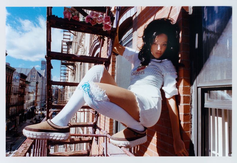 A woman in a playful pose on a fire escape outside a brick building with a city view, wearing a white top, white shorts, and platform shoes.