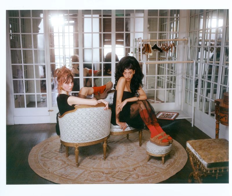 Two fashion models pose indoors seated on ornate chairs. One model has dark curly hair, the other has light pink hair. Wardrobe rack behind.