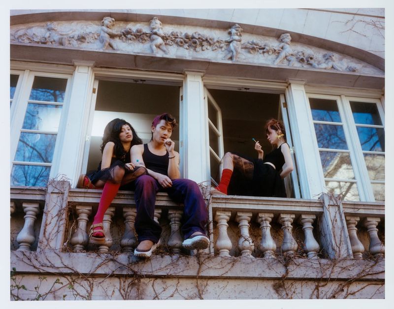 Three people sit in open windows of a building with classical architectural details, including vines, columns and decorative facade.