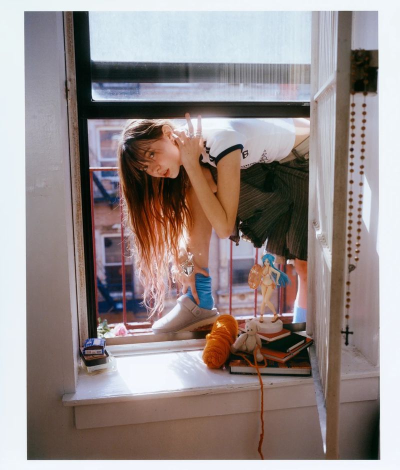 Young woman poses at an open window with decorative objects on the windowsill, showcasing casual fashion and an urban lifestyle.
