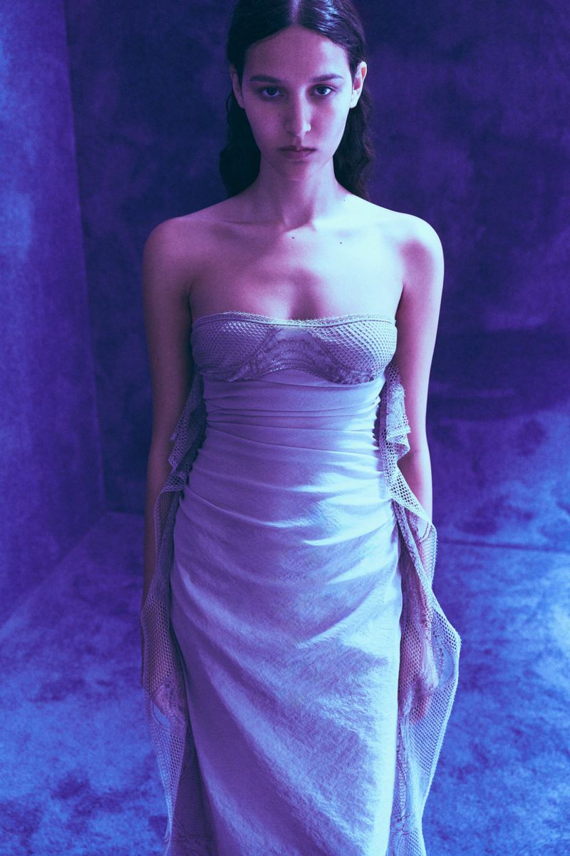 Portrait of an adult female fashion model posing in a floor-length strapless lilac gown with a textured background and studio lighting.