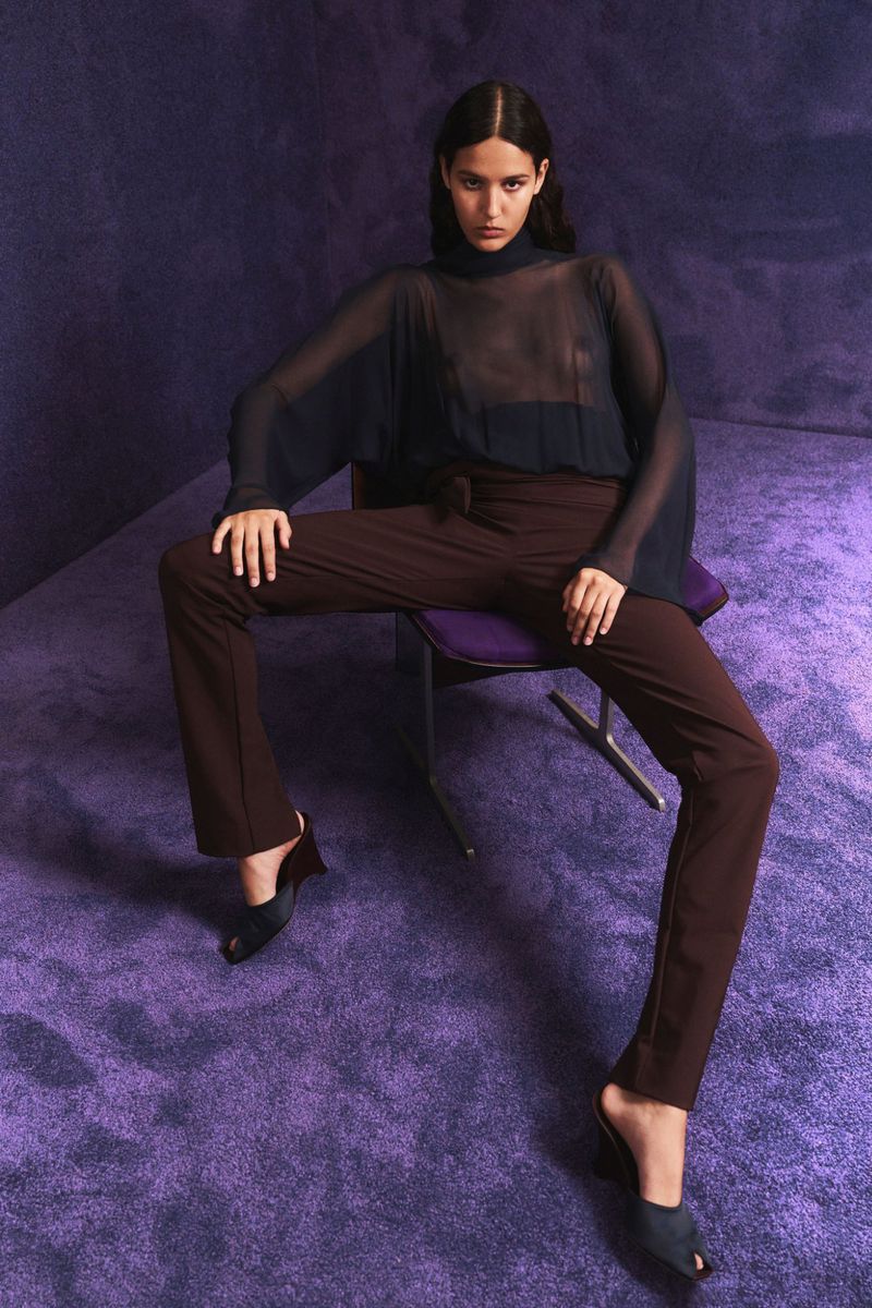A slender woman in a sheer top and brown pants sits in a photo studio on a chair in a full shot with a purple textured backdrop.