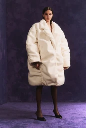 Model wearing an oversized white textured coat and black heels against a purple textured background.