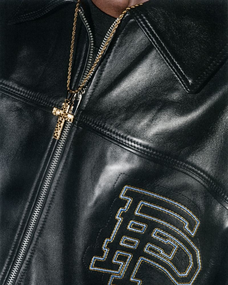 Close-up shot of a person wearing a black leather jacket with a gold cross necklace. The jacket features an embroidered design.