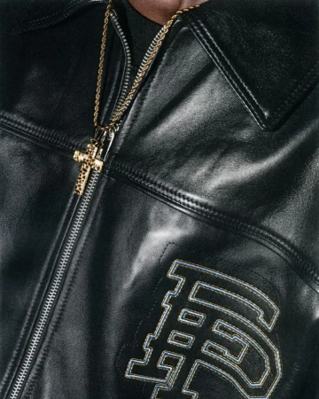 Close-up shot of a person wearing a black leather jacket with a gold cross necklace. The jacket features an embroidered design.