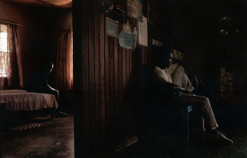 Dark, atmospheric interior scene featuring a man sitting in a chair in one room and another figure on a bed in the adjoining room.