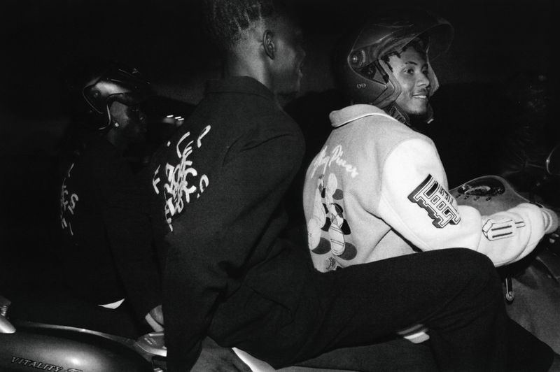 Two people riding a motorcycle at night, wearing helmets and graphic jackets, illuminated by dim lighting in an urban setting.