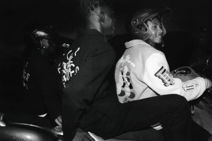Two people riding a motorcycle at night, wearing helmets and graphic jackets, illuminated by dim lighting in an urban setting.