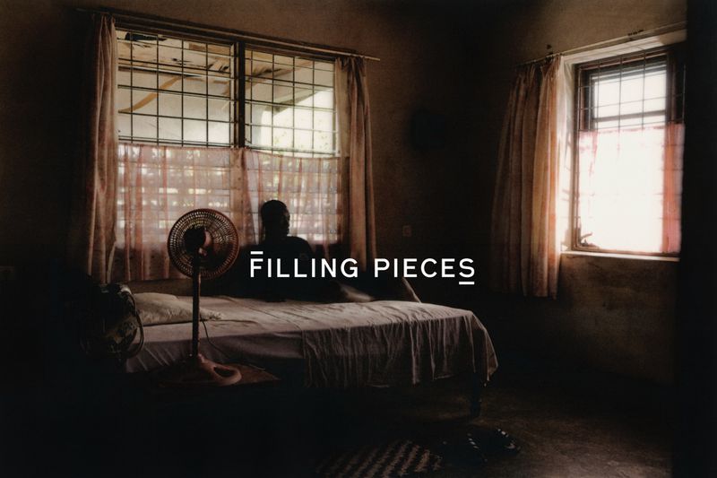 Dark interior scene with a silhouetted person on a bed, illuminated by window light, alongside a vintage fan and simple decor.