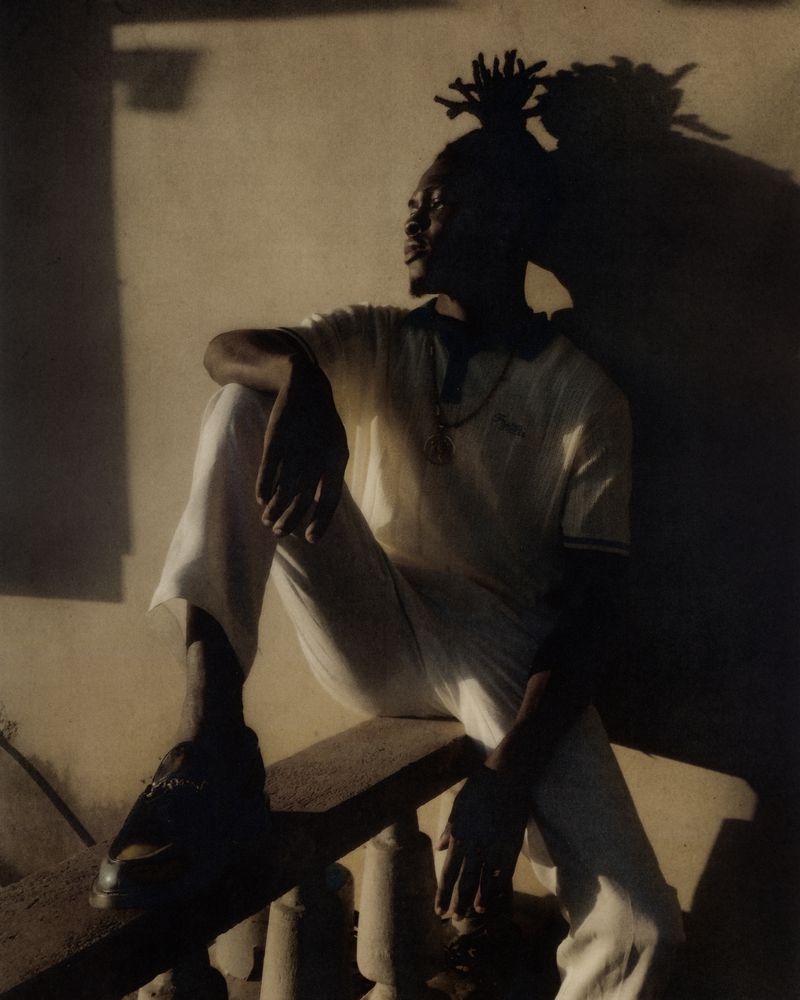 Man with dreadlocks sits outside in a white t-shirt and pants. Shadows add depth to the image. He looks off into the distance.