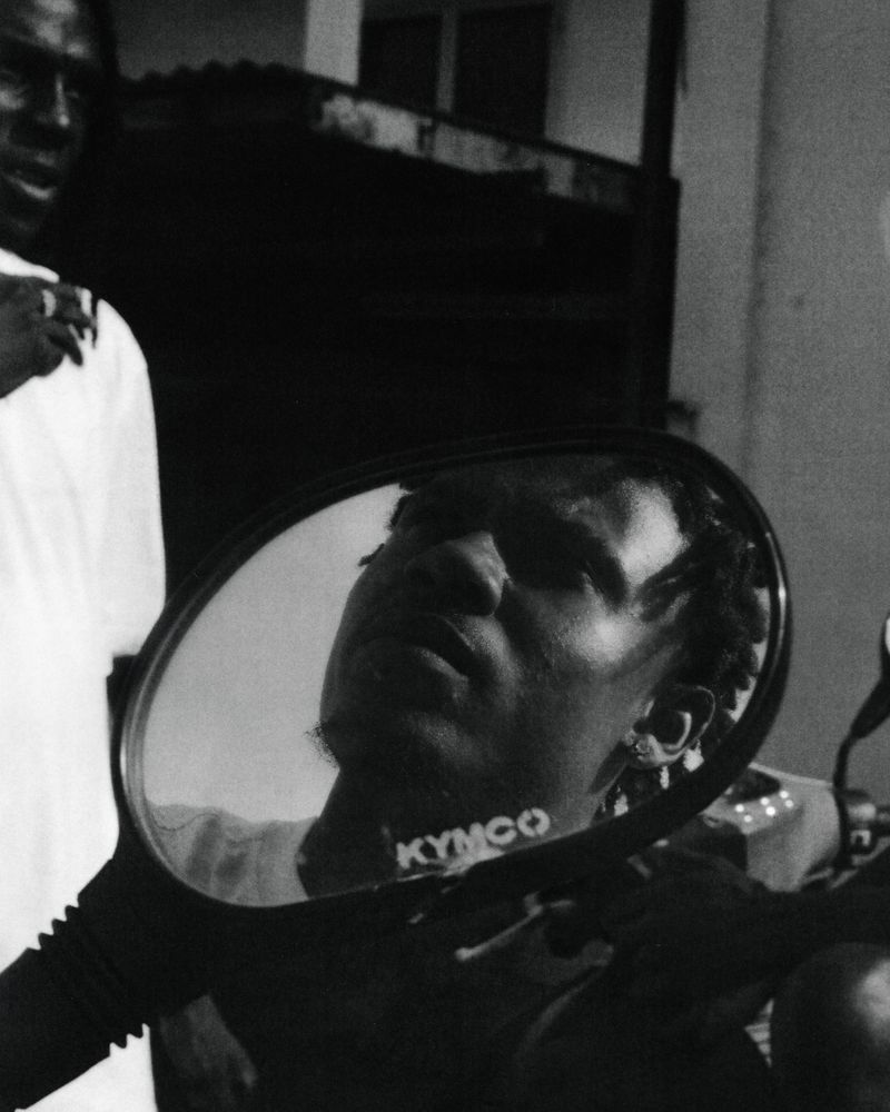 Monochrome portrait of a Black man reflected in the side mirror of a scooter in an urban setting, with another person visible in the background.