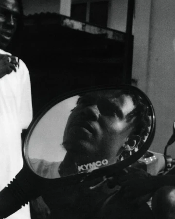 Monochrome portrait of a Black man reflected in the side mirror of a scooter in an urban setting, with another person visible in the background.