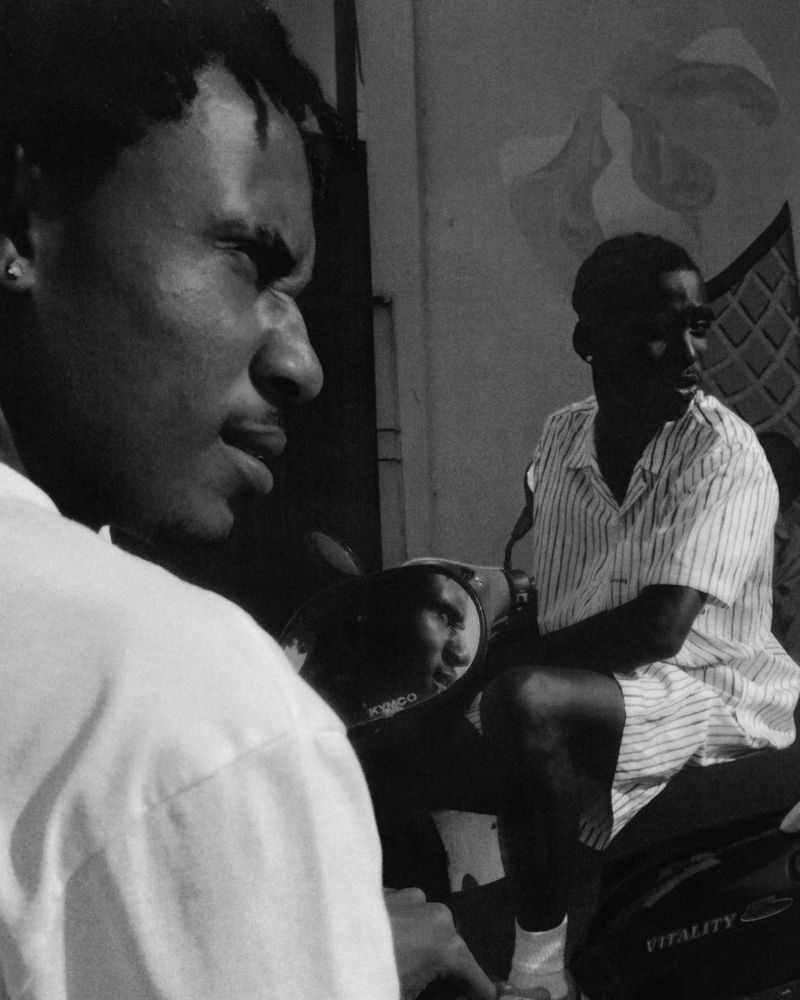 Monochrome medium shot of two adult males wearing collared shirts, one seated on a scooter, the other with a side profile expression.
