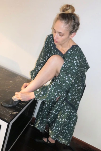 An adult woman sitting indoors, wearing a green floral dress and black shoes, appears to be putting on her shoe next to a black counter.