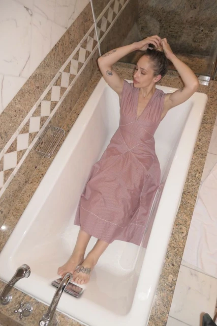 Woman in dusty rose dress relaxes in a white bathtub with her hands arranging her hair in an indoor bathroom setting.