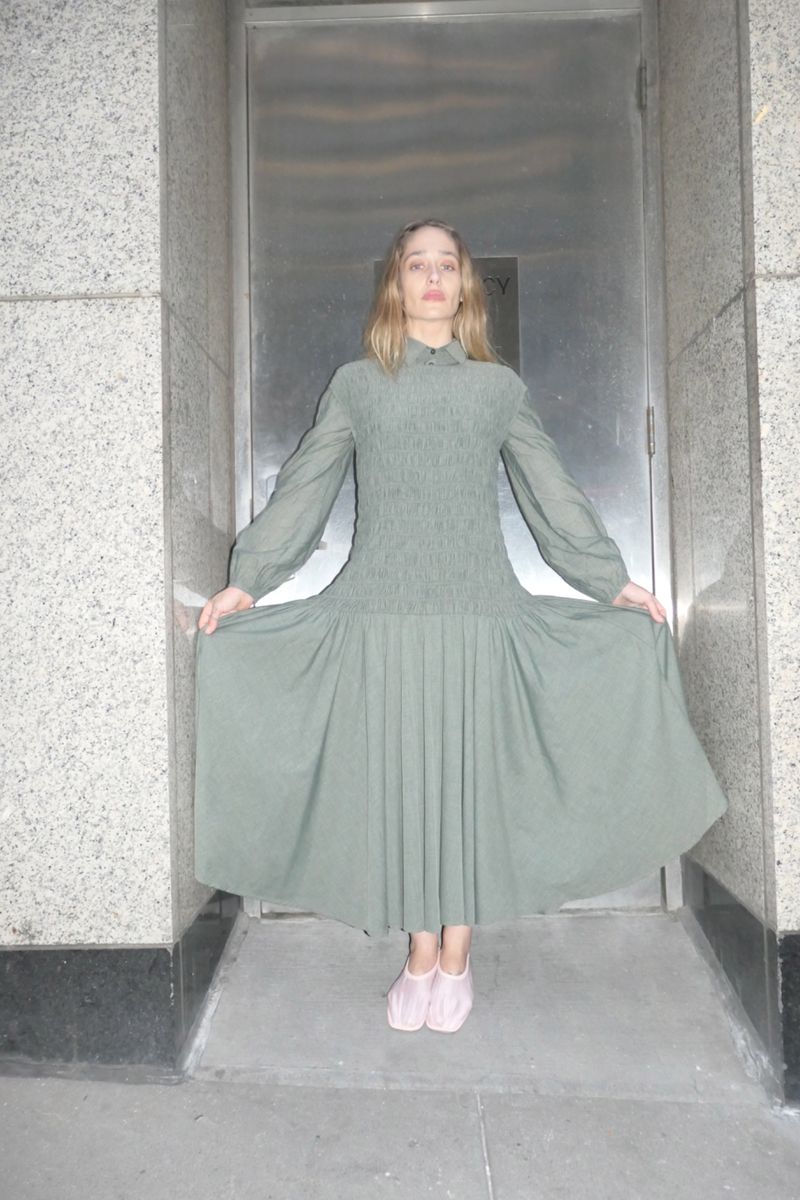 A fair-skinned young adult woman stands in a light green dress, holding the skirt edges, with a neutral expression in an indoor doorway.
