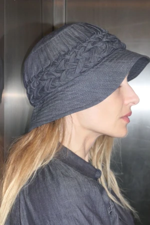 A woman in profile wears a dark grey bucket hat with a braided band and matching shirt against a reflective metal wall.