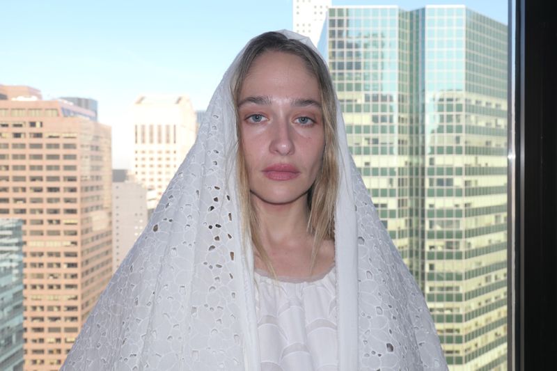Woman with blond hair poses in front of a city skyline, wearing a white lace veil and a white dress, in a medium shot.